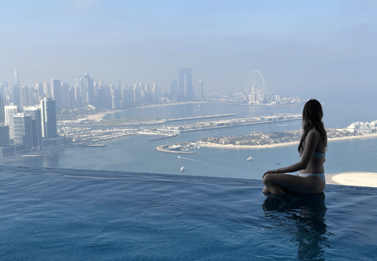 אישה יושבת על שפת בריכת אינסוף בבריכת אאורה בדובאי, עם נוף פנורמי של המרינה ואי הדקלים.