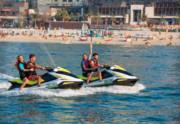 קבוצת אנשים רוכבת על אופנועי ים מול חופי דובאי, עם נוף של חוף ים ומבנים מודרניים מאחור.