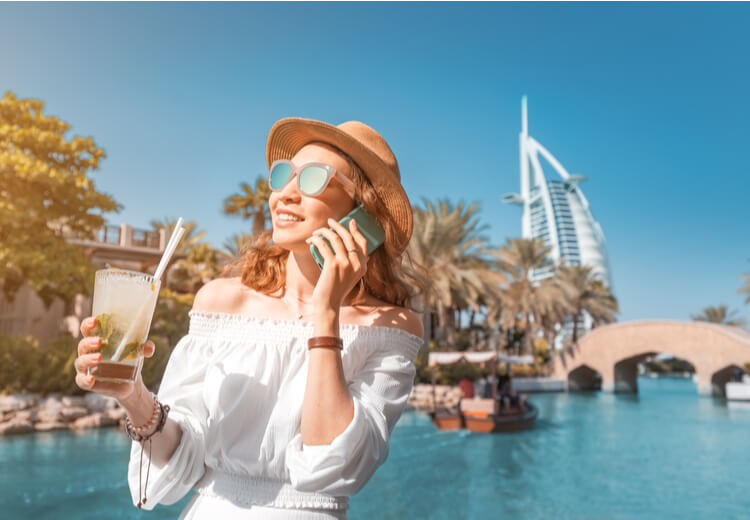 אישה בלבוש קיץ מדברת בטלפון ומחזיקה משקה קר ליד תעלה בדובאי, כשמלון בורג' אל ערב נראה ברקע ביום שמשי.
