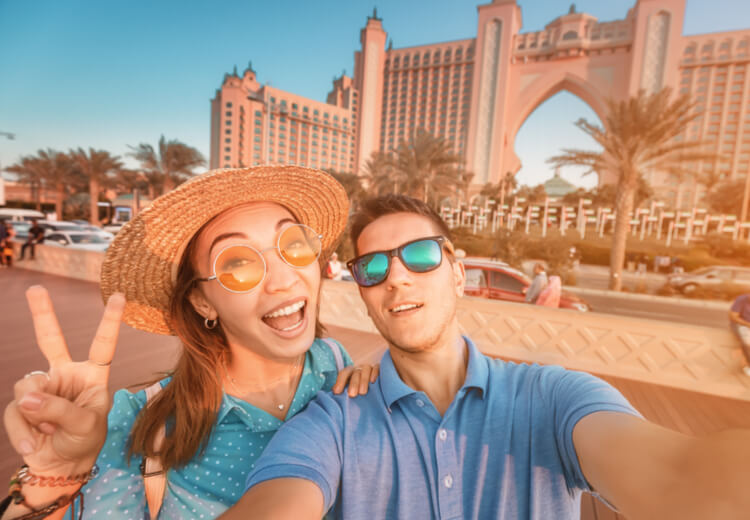 זוג תיירים מצטלם סלפי מול מלון אטלנטיס שבאי הדקל בדובאי, ביום שמשי עם אווירה שמחה ונינוחה.